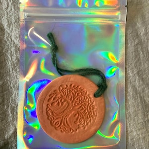 May include: A circular, terracotta-coloured ornament featuring a tree of life design, attached to a green string. The ornament is enclosed in a holographic, iridescent plastic bag with a zip closure.