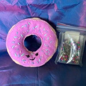 May include: A pink felt donut with sprinkles and a smiling face beside a clear plastic bag filled with colourful sewing pins. The donut has a light tan edge and blue and green sprinkles.