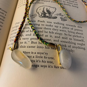May include: A close-up of a book with a Winnie the Pooh illustration. A colorful braided bookmark with two white stone charms hangs from the page. The text on the page reads "Here is a myst'ry out a little fir-tree. Ays it's his tree, anga says it's her tre..."