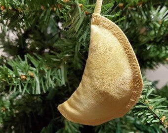 May include: A pale yellow crescent moon-shaped ornament made of fabric hangs from a white ribbon on a green Christmas tree.