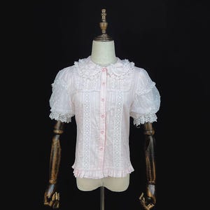 May include: A light pink blouse with short, puffy sleeves and a Peter Pan collar. The blouse features white lace detailing down the front and around the collar and sleeves. The sleeves have a sheer, dotted fabric. The blouse is buttoned up the front.