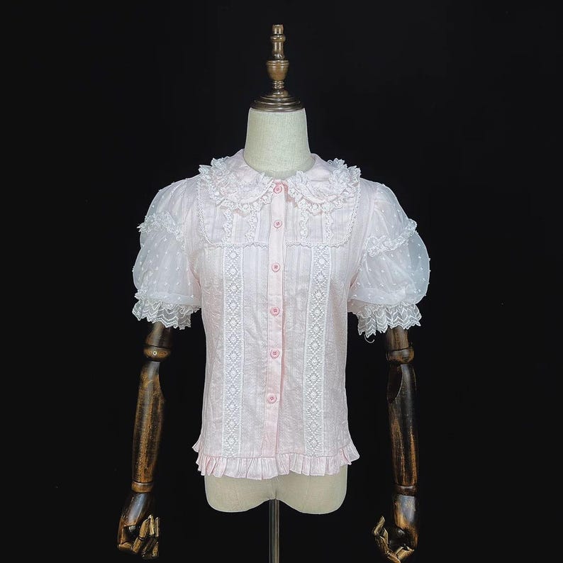 May include: A light pink blouse with short, puffy sleeves and a Peter Pan collar. The blouse features white lace detailing down the front and around the collar and sleeves. The sleeves have a sheer, dotted fabric. The blouse is buttoned up the front.