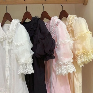 May include: Four long-sleeved blouses in white, black, pink, and yellow, hanging on wooden hangers. Each blouse features lace trim on the collar, sleeves, and hem. The white and pink blouses have a dotted pattern on the sleeves.