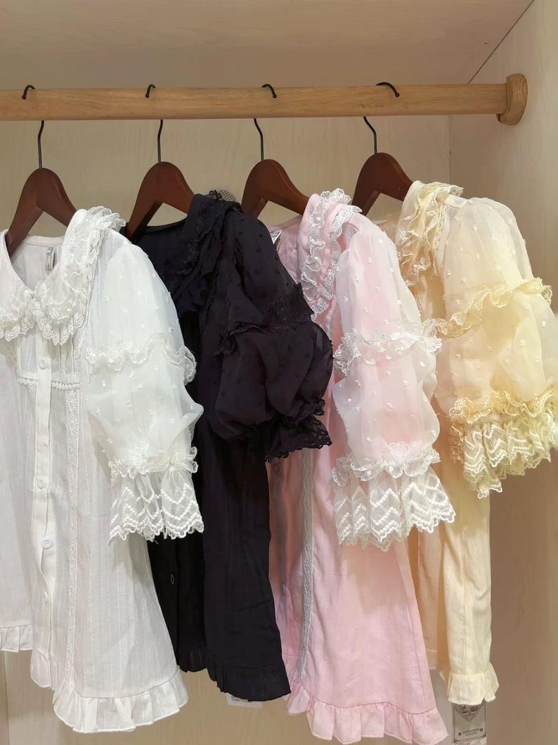 May include: Four long-sleeved blouses in white, black, pink, and yellow, hanging on wooden hangers. Each blouse features lace trim on the collar, sleeves, and hem. The white and pink blouses have a dotted pattern on the sleeves.