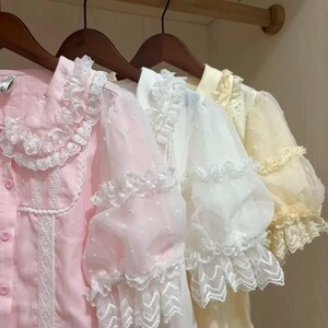May include: Three blouses in pastel colors hang on wooden hangers. The blouses have lace trim and ruffled sleeves. The first blouse is pink, the second is white, and the third is yellow.