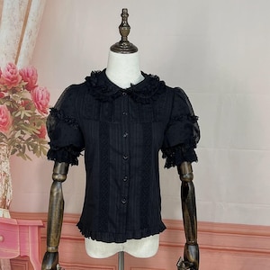 May include: Black short-sleeved blouse with a ruffled collar and cuffs. The blouse features a button-down front, vertical stripes, and lace detailing. The sleeves are sheer with a dotted pattern. The blouse is displayed on a mannequin.