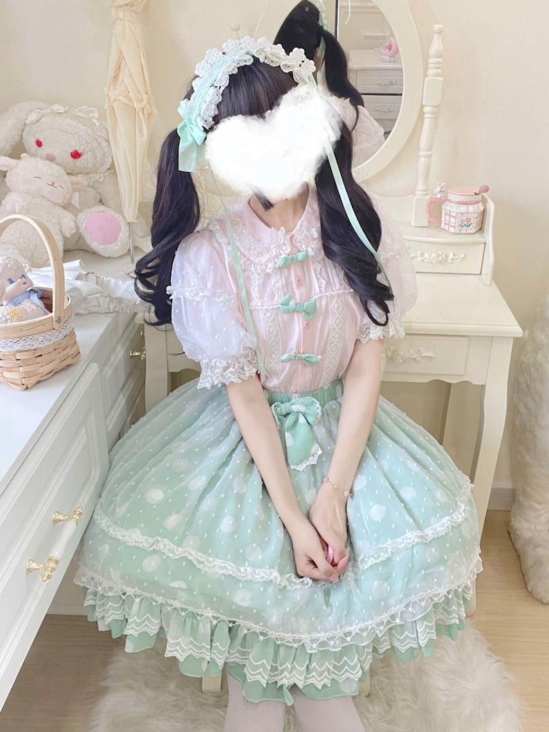 May include: A pastel pink and mint green Lolita dress. The dress features a white lace collar, puff sleeves, and a mint green skirt with white polka dots. The outfit is accessorized with a matching headband and bows.