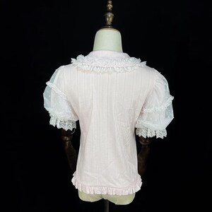 May include: A light pink blouse with a ruffled collar and hem. The short sleeves are sheer white with a polka dot pattern and lace trim. The blouse is displayed on a mannequin against a black background.