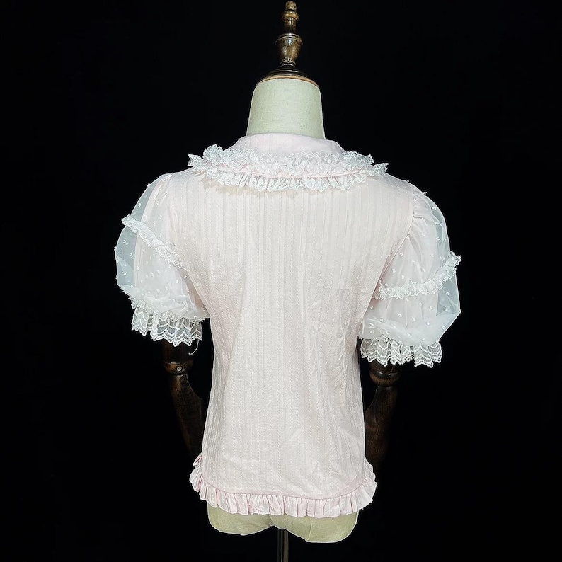May include: A light pink blouse with a ruffled collar and hem. The short sleeves are sheer white with a polka dot pattern and lace trim. The blouse is displayed on a mannequin against a black background.