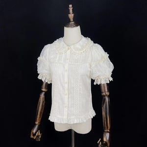 May include: White short-sleeved blouse with a Peter Pan collar and button-down front. The blouse features lace and ruffle detailing on the collar, sleeves, and hem. The sleeves are puffed with a dotted sheer fabric. The blouse is displayed on a mannequin.