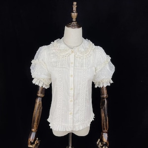 May include: White blouse with short, sheer, dotted sleeves and a Peter Pan collar. The blouse features lace trim on the sleeves, collar, and front panel. It has a button-down front and a ruffled hem, creating a vintage aesthetic.