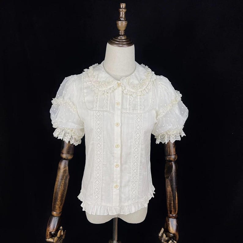 May include: White blouse with short, sheer, dotted sleeves and a Peter Pan collar. The blouse features lace trim on the sleeves, collar, and front panel. It has a button-down front and a ruffled hem, creating a vintage aesthetic.