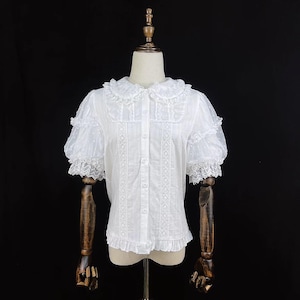 May include: White blouse with a Peter Pan collar, short puff sleeves, and decorative lace trim. The blouse features a button-down front and a ruffled hem. The fabric has a subtle dotted pattern. The blouse is displayed on a mannequin.