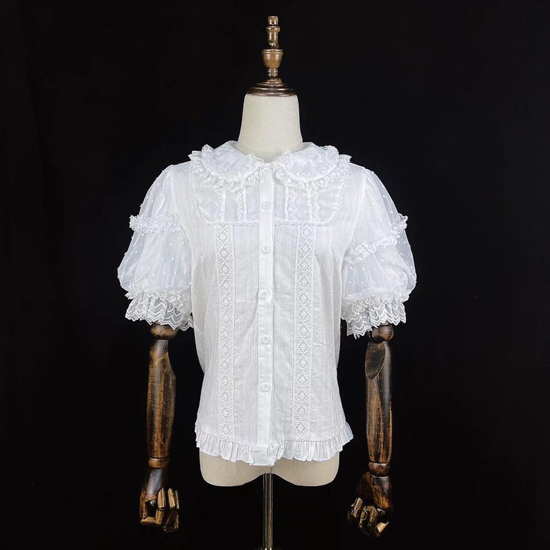 May include: White blouse with a Peter Pan collar, short puff sleeves, and decorative lace trim. The blouse features a button-down front and a ruffled hem. The fabric has a subtle dotted pattern. The blouse is displayed on a mannequin.