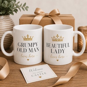Könnte beinhalten: Zwei weiße Keramikbecher mit goldenem Text und Kronen. Auf einem Becher steht "GRUMPY OLD MAN lives here" und auf dem anderen "BEAUTIFUL LADY lives here". Eine Karte lautet "Welcome to your castle". Eine Geschenkbox und ein Band sind im Hintergrund.
