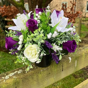 May include: A floral arrangement in a black pot. The arrangement features white lilies, purple roses, and white and purple accent flowers, with green foliage. The arrangement is set on a wooden fence.