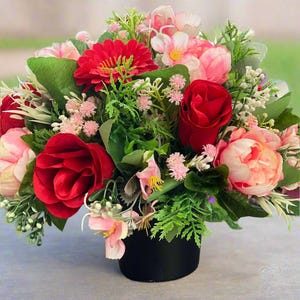 Pode incluir: Um arranjo de flores artificiais em um vaso preto, com rosas vermelhas, gerberas e outras flores em tons de rosa e verde.