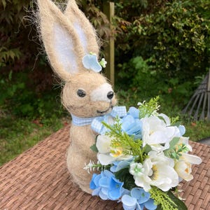 May include: A decorative Easter bunny figurine made of tan woven material, with long ears and a blue gingham bow. The bunny holds a bouquet of white and blue flowers with green foliage, perfect for spring decor.