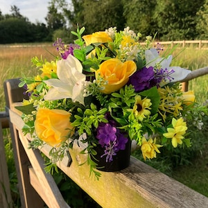 Pode incluir: Um arranjo de flores artificiais amarelas e roxas em um vaso preto. O arranjo inclui rosas amarelas, lírios brancos e flores roxas.