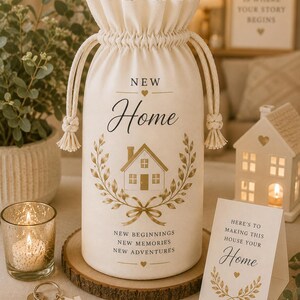 Op de afbeelding: Een crèmekleurige wijnfleszak met een trekkoord, met de woorden "New Home" en een huisillustratie in goud. De tas bevat ook de zinnen "New Beginnings, New Memories, New Adventures."