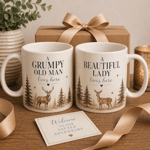 Könnte beinhalten: Zwei weiße Keramikbecher mit den Aufschriften "A GRUMPY OLD MAN lives here" und "A BEAUTIFUL LADY lives here". Jeder Becher zeigt ein Reh und eine Waldszene. Eine kleine Karte trägt die Aufschrift "Welcome TO OUR LITTLE ADVENTURE".
