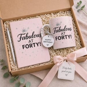 May include: A gift box containing a light pink notebook and a matching ceramic mug, both printed with the text Fabulous AT FORTY! in black and silver. A silver pen, a Happy Birthday tag, and a pink ribbon with a Forty looks good on you tag.
