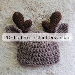 May include: A brown crocheted reindeer hat with large antlers. The hat is on a white fuzzy surface. The text "PDF Pattern | Instant Download" is on the image.