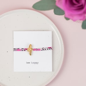 May include: A white card with the text "bee happy" printed on it. A pink and white patterned ribbon is tied around the card with a gold bee charm in the centre.
