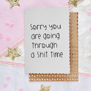 May include: A white card with a scalloped edge, featuring the text "Sorry you are going through a shit time" in black. The card is on a brown envelope with gold star patterns. Pink confetti and gold glitter stars decorate the background.