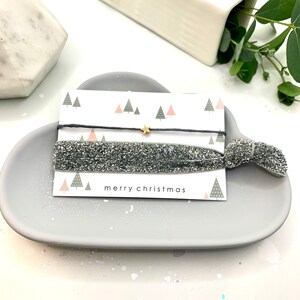 May include: A grey, oval dish displays two hair ties and a card. One hair tie is black with a gold star charm, the other is silver glitter. The card reads "merry christmas" and features a Christmas tree design.
