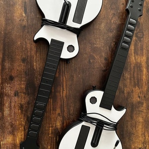 Peut inclure: Deux contrôleurs de jeu en forme de guitare, blancs et noirs. Les contrôleurs ont des manches et des touches noirs, avec le logo Gibson sur l'un d'eux. Les corps sont blancs avec des accents et des boutons noirs. Les contrôleurs sont reliés par des câbles noirs.