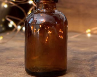 May include: A small, cylindrical amber glass bottle with a short, narrow neck and a wide, flat rim. The glass is translucent and brown, featuring a smooth, rounded body that tapers slightly toward the top opening, creating a classic apothecary style.