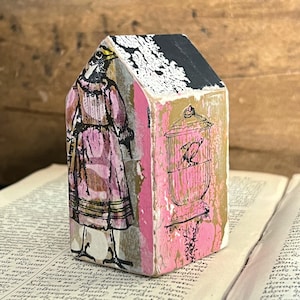 May include: A small house-shaped wooden block featuring a collage design. One side displays a bird in a pink dress, while the other side shows a black ink illustration of a bird inside a cage. The roof is painted black with white textured details.
