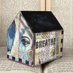 May include: A small house-shaped wooden block featuring a black roof and a collage design on the sides. The artwork includes a detailed human eye, vintage text, and the word BREATHE in bold black letters, accented with blue and cream tones.