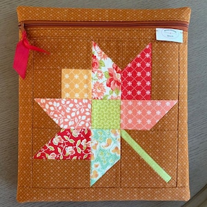 May include: A square fabric zipper pouch featuring a quilted maple leaf design in shades of orange, red, yellow, and green. The background fabric is a dark orange with a small white geometric pattern. A red ribbon is attached to the zipper pull.