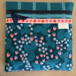 May include: A square, zippered pouch with a dark teal strawberry pattern. The pouch has a black zipper, a navy blue ribbon, and a band of pink strawberries. The top section has a teal and white dotted pattern.