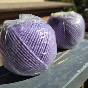 May include: Two spherical balls of light purple yarn, each individually wrapped in clear, crinkled plastic film. The yarn features a twisted, multi-ply texture and a consistent lavender hue throughout the wound fibers of both round skeins.