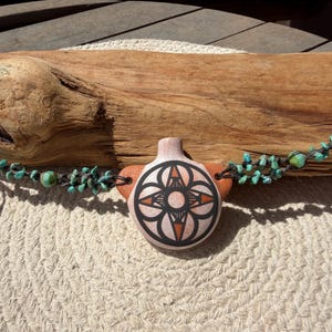 May include: A round, terracotta-colored ceramic pendant featuring a black and orange geometric star pattern. The pendant is suspended on a dark cord adorned with small, irregular turquoise-colored stone beads on both sides of the central piece.