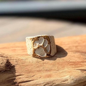 May include: A ceramic ring featuring a speckled off-white finish and a recessed paw print design. The ring has a wide band construction with a textured surface and dark brown accents highlighting the paw print impression on the front face.
