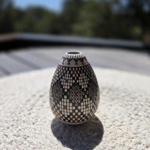 May include: A decorative, patterned vase with a narrow neck and a rounded body. The vase features a geometric design in shades of brown, black, and white. The vase is sitting on a textured, off-white surface, with a blurred background of trees and sky.