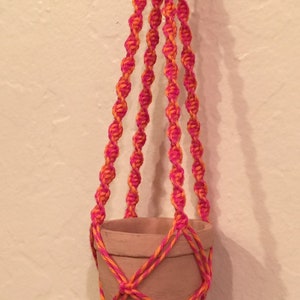 May include: A handmade macrame plant hanger in vibrant shades of orange and pink. The plant holder is designed to suspend a small terracotta pot. The decorative hanger features a knotted design and a tassel at the bottom.