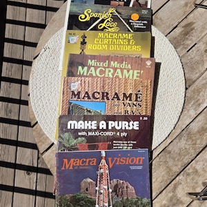 May include: A stack of vintage macrame books, including titles like "Purses Round the World" and "Macra Vision." The covers feature images of macrame projects, with text in various fonts and colors. The books are on a wooden table.