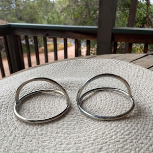 May include: Two silver napkin rings with a curved handle design. The rings are circular and sit on a light beige woven placemat. The background shows a wooden deck and trees.