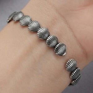 May include: A silver cuff bracelet featuring a repeating pattern of small, detailed seashell motifs. The metal has an oxidized finish that highlights the ridges and textures of each individual shell design along the open-ended circular band.