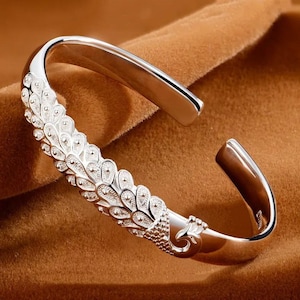 May include: A silver cuff bracelet with a peacock feather design. The bracelet features a detailed pattern of feathers and a peacock head at one end. The metal has a polished, reflective surface, and is set against a brown background.