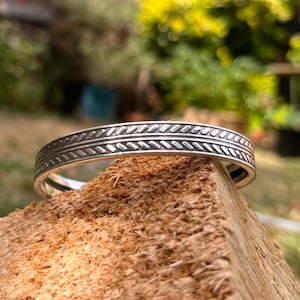 Pode incluir: Uma pulseira de metal em tom prateado com um padrão de fila dupla de linhas diagonais repetitivas gravadas. A pulseira tem um acabamento liso e polido e um design circular aberto e contínuo com largura uniforme em toda a sua extensão.
