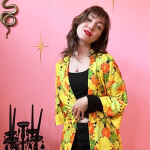 May include: A yellow floral kimono with orange and green accents. The kimono is open in the front and tied at the waist. The background is a pink wall with gold starburst decorations.