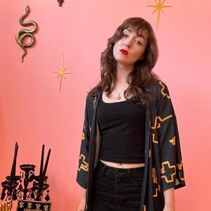 May include: Black kimono with a gold geometric pattern. The open-front kimono is worn over a black top and black pants. The background is a pink wall with gold starburst decorations and a gold snake.