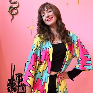 May include: A colorful kimono-style jacket with a vibrant pattern of dripping shapes in pink, yellow, and blue, outlined in black. The jacket is open, revealing a black top and long-sleeved shirt. The background is a solid pink wall with gold star decorations.