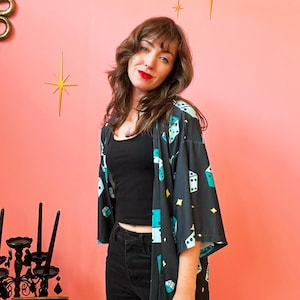 May include: A black kimono-style robe with a dice pattern in teal, white, and yellow. The robe is open, revealing a black top and black jeans. The background is a solid pink wall with gold starburst decorations.